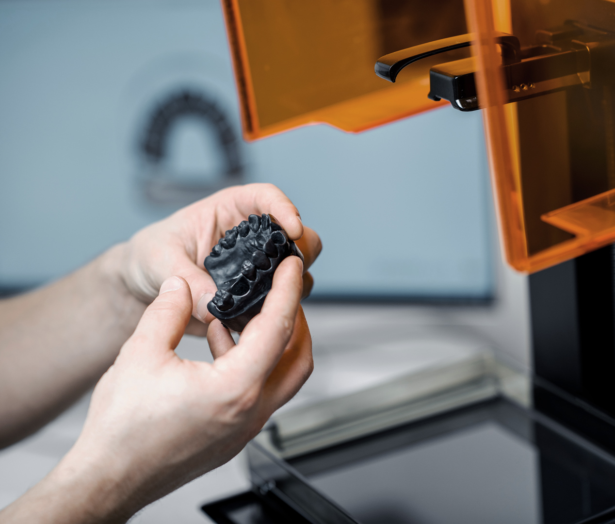 Hands holding a 3D-printed dental model near a 3D printer, with the printer's orange cover partially open in Des Moines IA.