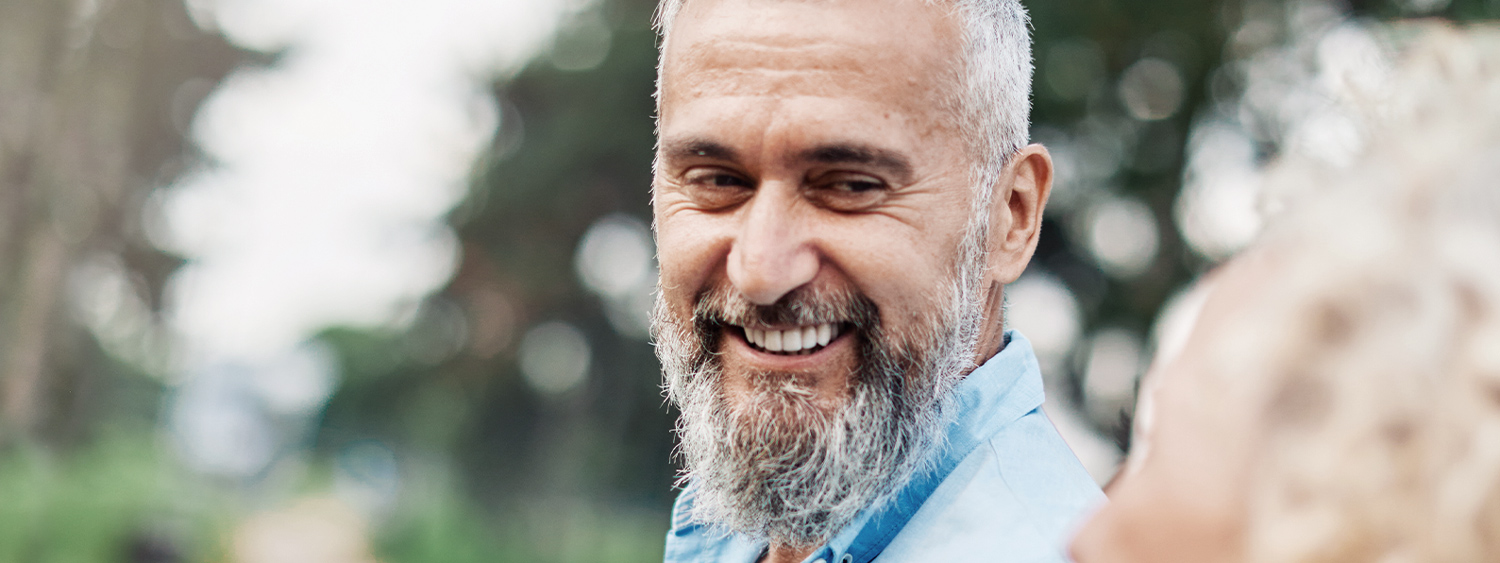 A bearded man smiling warmly while conversing outdoors, with greenery in the background in Des Moines IA. mobile